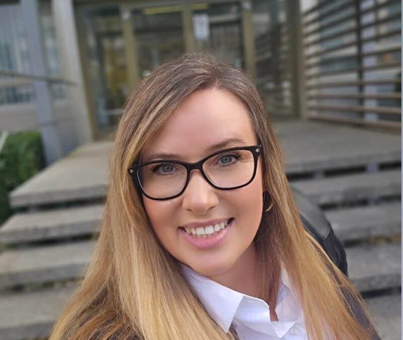 Frau mit langen Haaren und Brille steht vor dem Eingang eines Gebäudes der IHK.